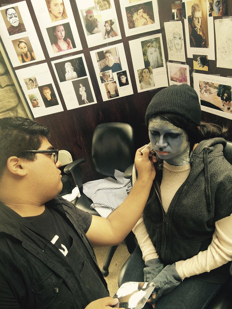 一名黑发男学生给一名戴着黑色无边帽的学生画上了怪物般的灰色角色妆容. 她们身后的墙上挂着化妆照.